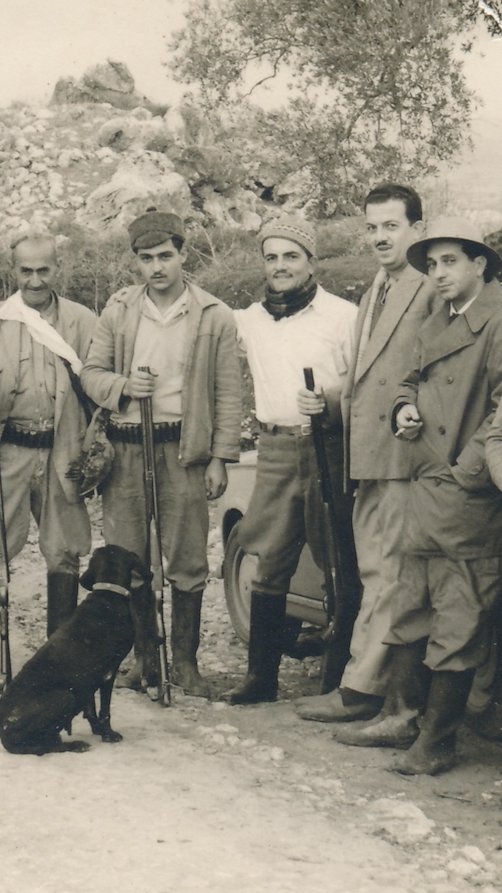 Black and white photo of five men standing with rifles and a dog, posing outdoors in a rocky landscape.